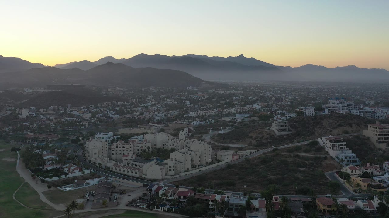 Hilltop view over pale buildings, roads, hills, and a mountain horizon at morning light.