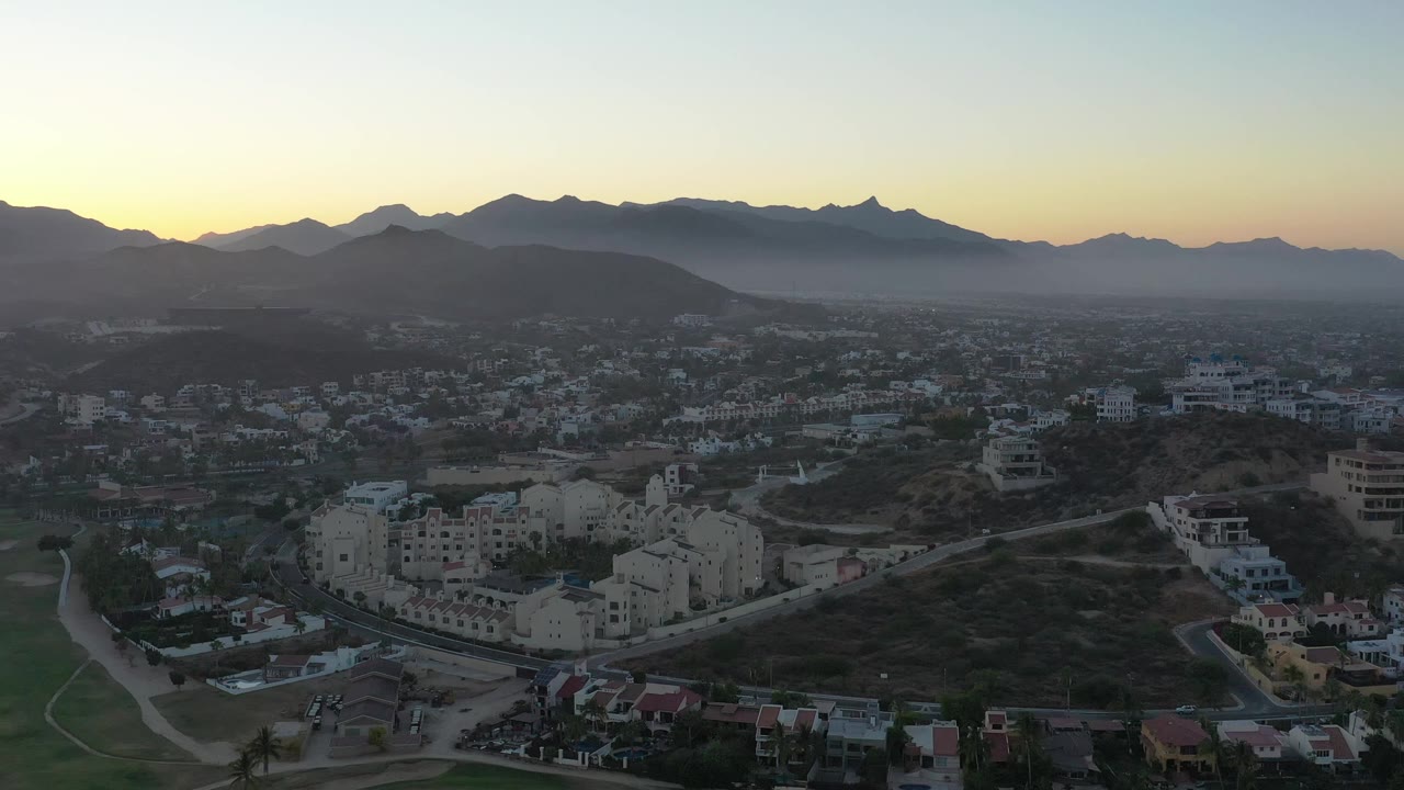 San Jose del Cabo, Mexico appears from a high vantage with white and beige buildings in the lower frame, layered hills behind them, a jagged ridge line, morning haze, and no visible people.