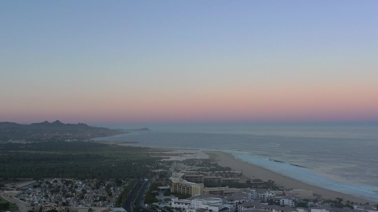 Wide aerial coastline with beach, ocean, hills, buildings, and pastel sky.