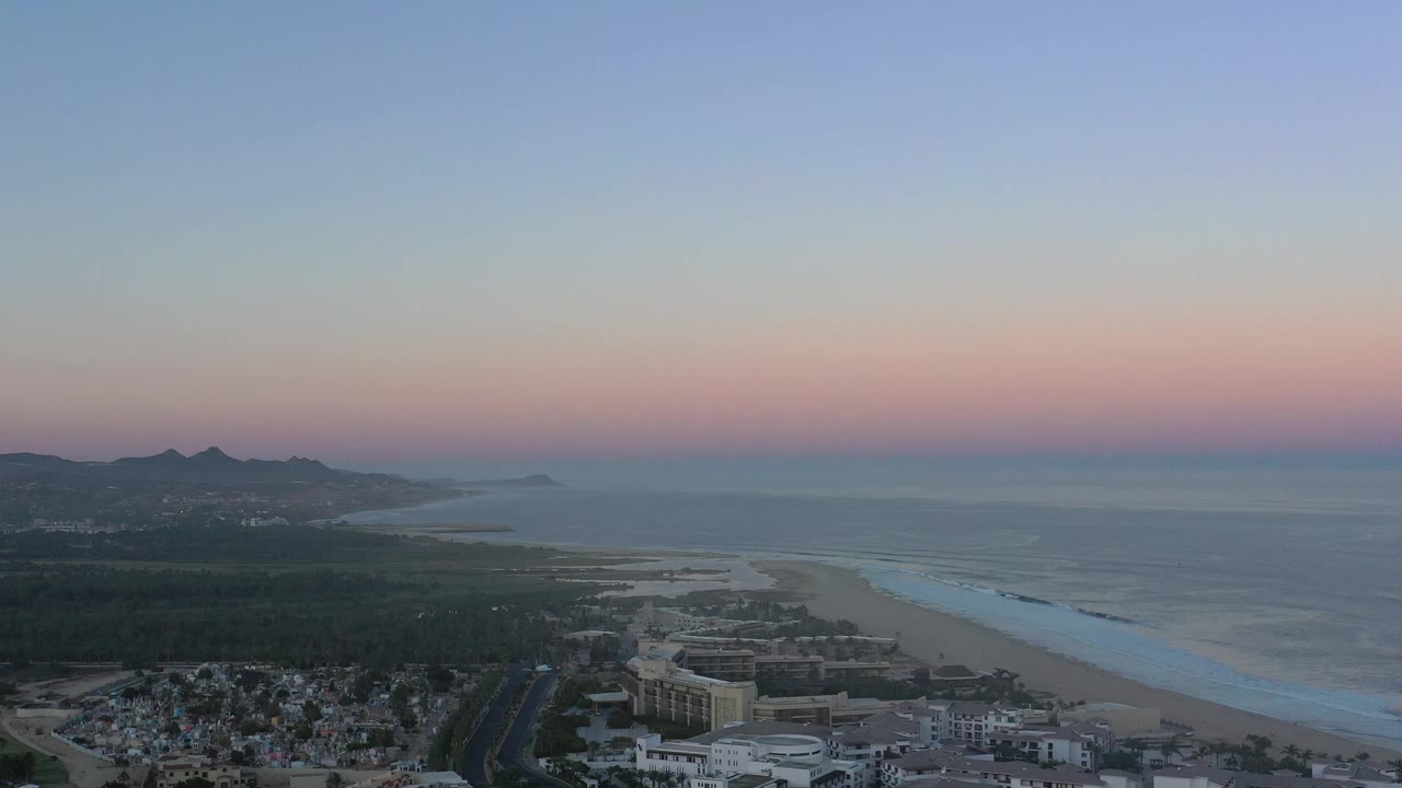 San Jose del Cabo coastline from above, with the surf line stretching along the beach, hills silhouetted on the left, and no visible people.