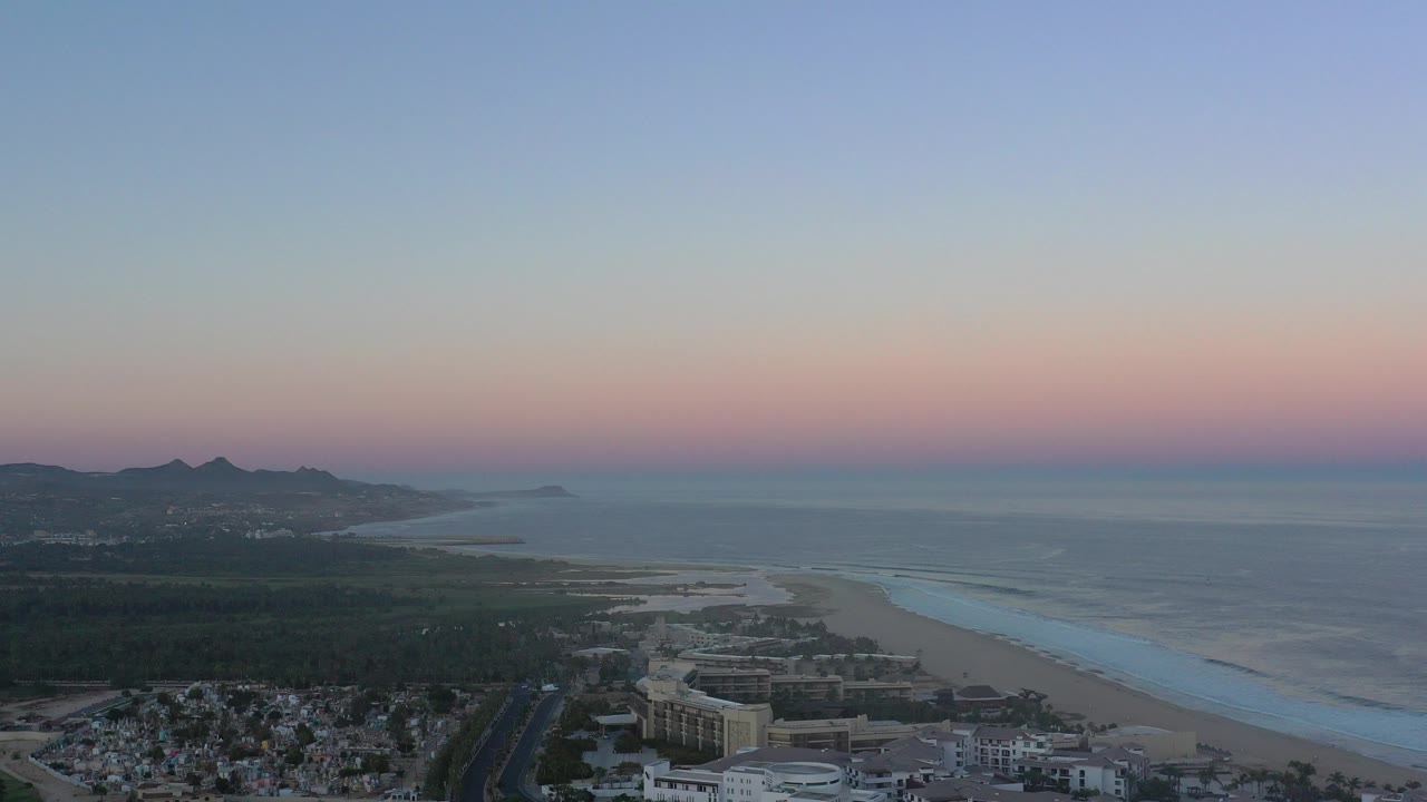 Aerial view of coastal houses, beach, sea, distant hills, and pastel sunset sky.