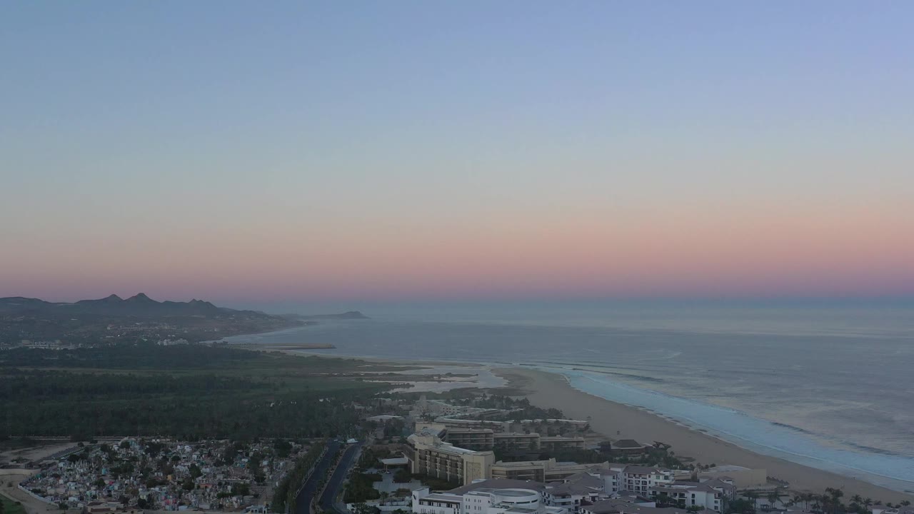 High view over San Jose del Cabo showing a darkening residential foreground, a road leading toward the beach, pale surf, open water, distant hills, and no visible people.