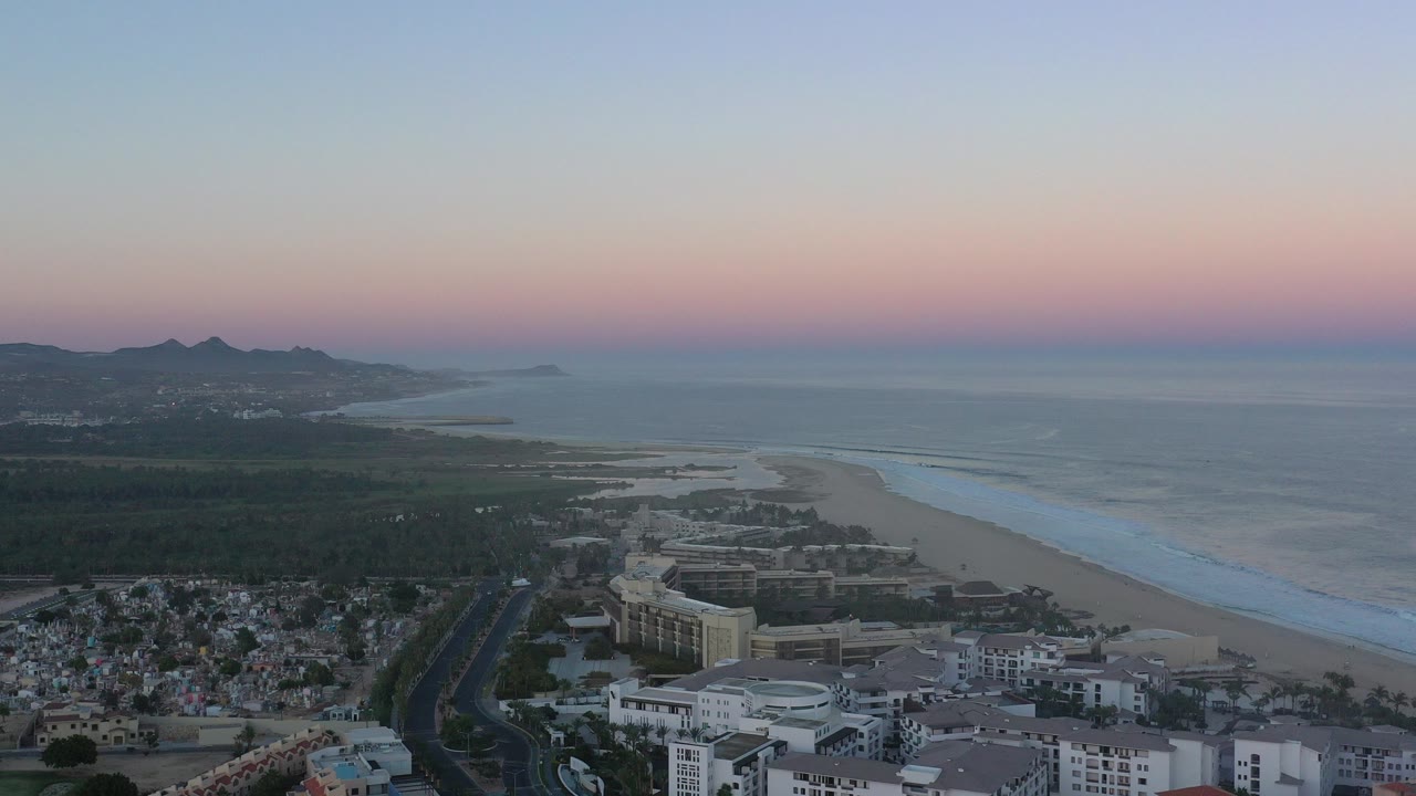 High aerial view over San Jose del Cabo, Mexico, with town buildings below, a sandy beach and surf line to the right, distant hills on the horizon, and no visible people.