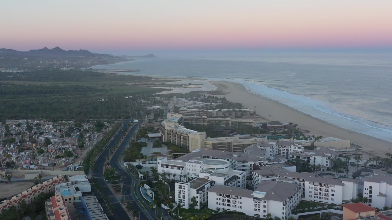 Aerial view over San Jose del Cabo with white buildings in the foreground, a road and residential area to the left, beach and sea to the right, mountains in the distance, and no visible people.