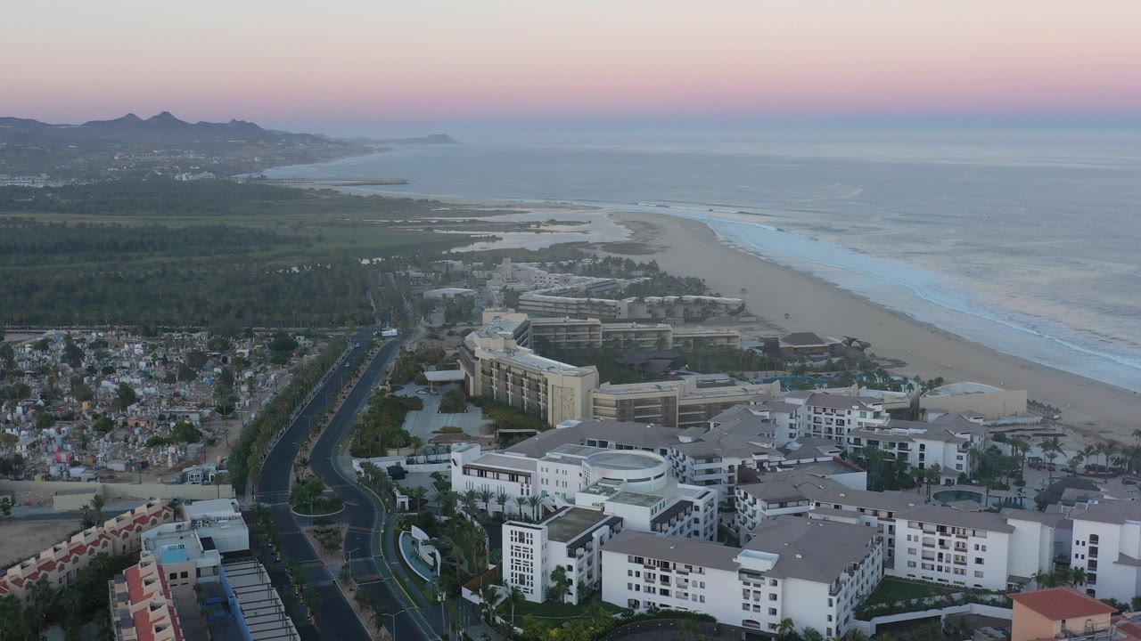 Aerial coastal view with white buildings, neighborhood blocks, a road, beach, ocean, and mountains under pastel sky.