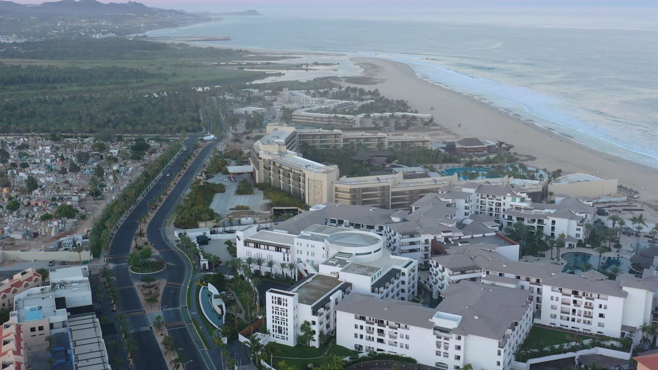 Opening aerial frame over San Jose del Cabo showing resort buildings in the foreground, a curved road at left, beach and ocean at right, distant hills, and no visible people.