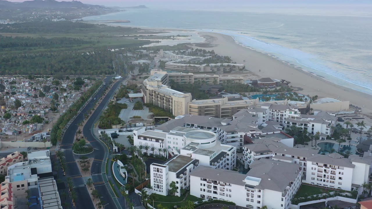Aerial view of San Jose del Cabo with white hotel buildings lower in frame, a divided road running inland, beach and pale surf to the right, and no visible people.