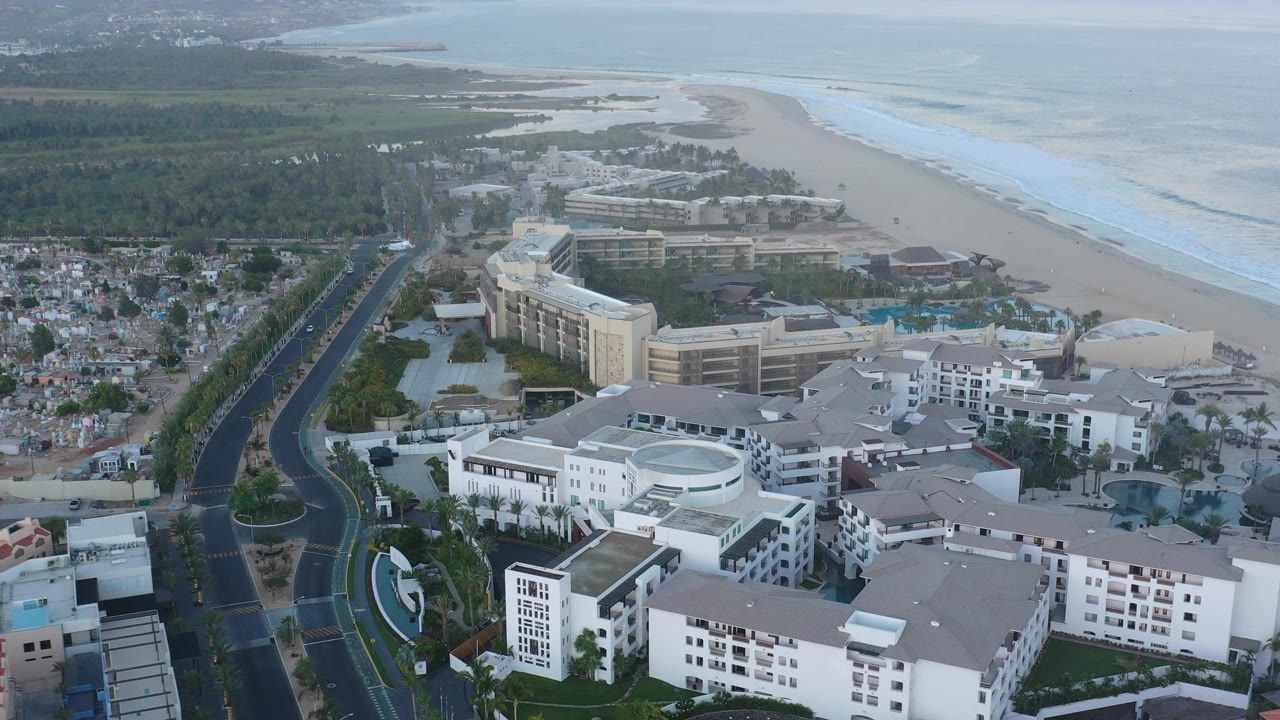 Resort area beside a beach with road, pools, palms, ocean, and hills.
