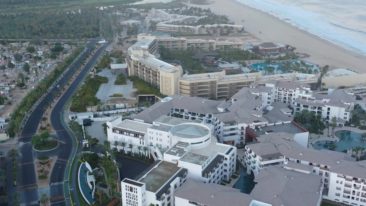 A slightly later aerial frame in San Jose del Cabo shows white resort blocks in the foreground, a dark curved road at left, blue pools near the beach, ocean surf, and no visible people.
