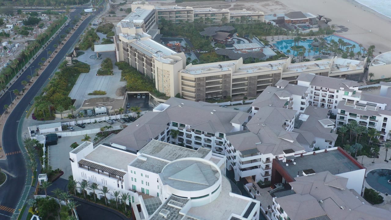 High aerial frame over San Jose Del Cabo resort buildings, with a palm-lined road at left, blue pools near the upper right, a sandy beach edge beyond, and no visible people.