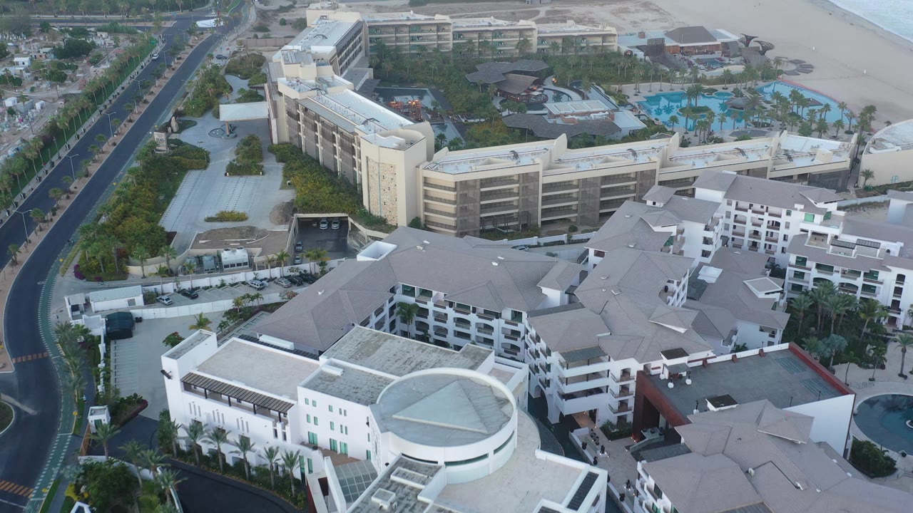 Aerial view in San Jose Del Cabo showing a curved white building in the lower foreground, beige hotel blocks in the midground, blue pools in the background, and no visible people.
