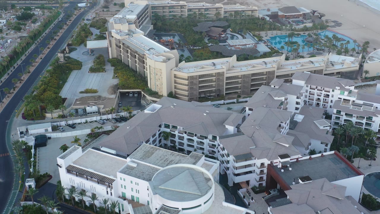 Drone view across white resort roofs, roads, pools, and beach.