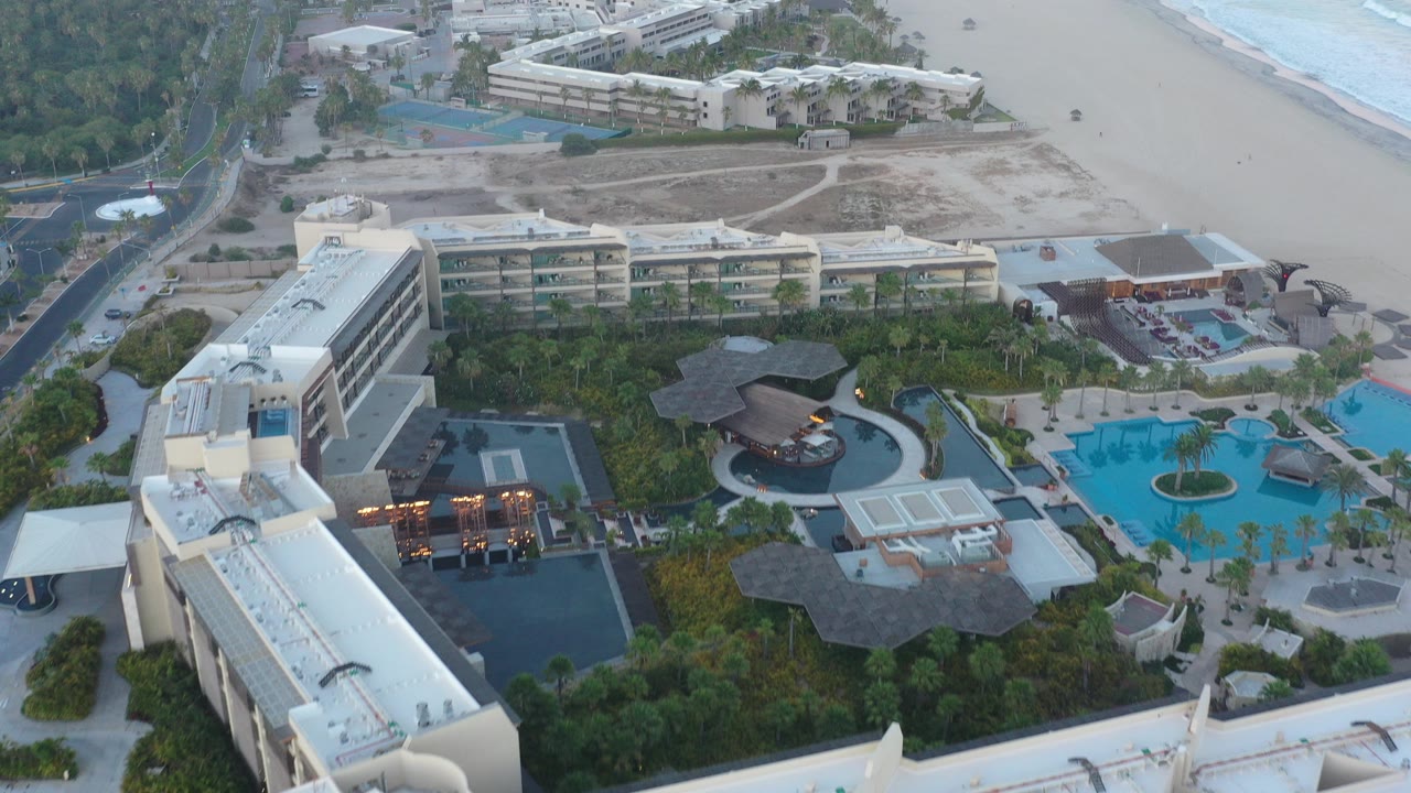 Aerial view of beachfront resort buildings, pools, palm landscaping, beach, and surf.