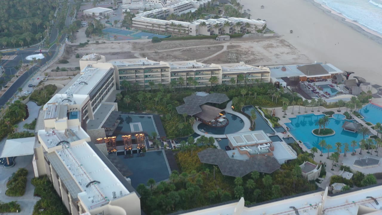 High aerial frame over San Jose Del Cabo resort grounds with a large white building in the lower left, pool courtyards near center, road at left, and beach with surf beyond.