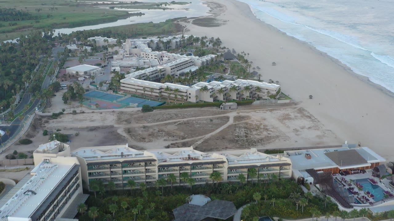 A high aerial frame over San Jose del Cabo shows beachfront buildings and tennis courts beside a wide sandy beach, pale surf, and green wetland channels, with no visible people.