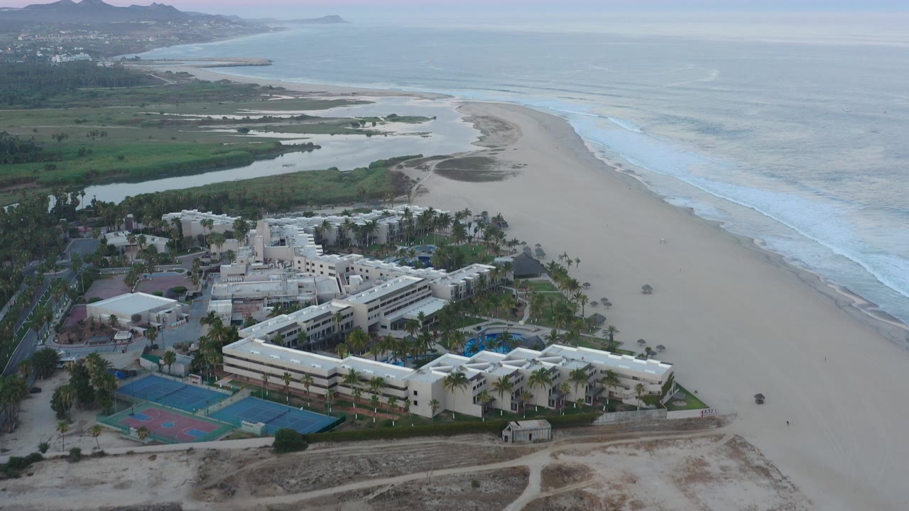 Aerial view in San Jose del Cabo showing a white modern building complex, blue courts, palms, empty beach, breaking surf, wetlands, distant hills, and no visible people.