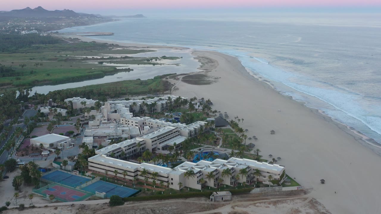 Wide drone view of a beachfront complex with surf and distant coastline.