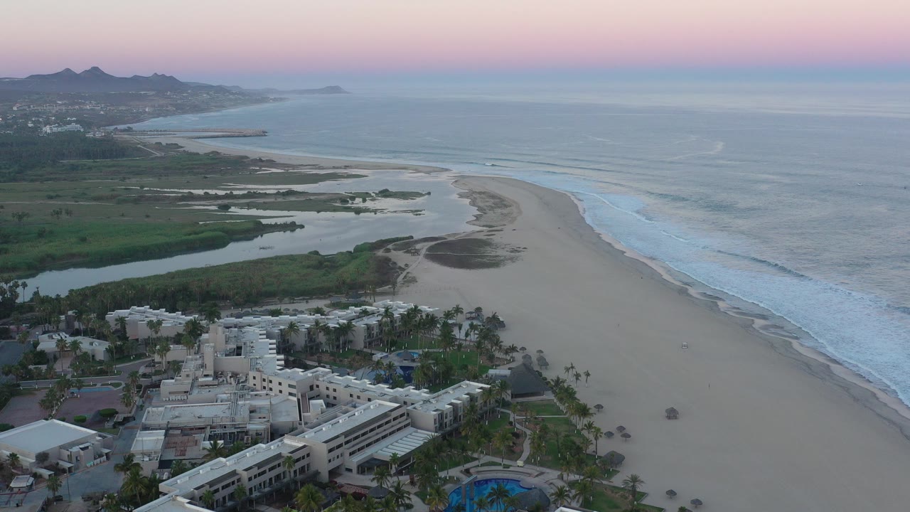 Aerial view of resort buildings beside a sandy beach and ocean surf at sunset.