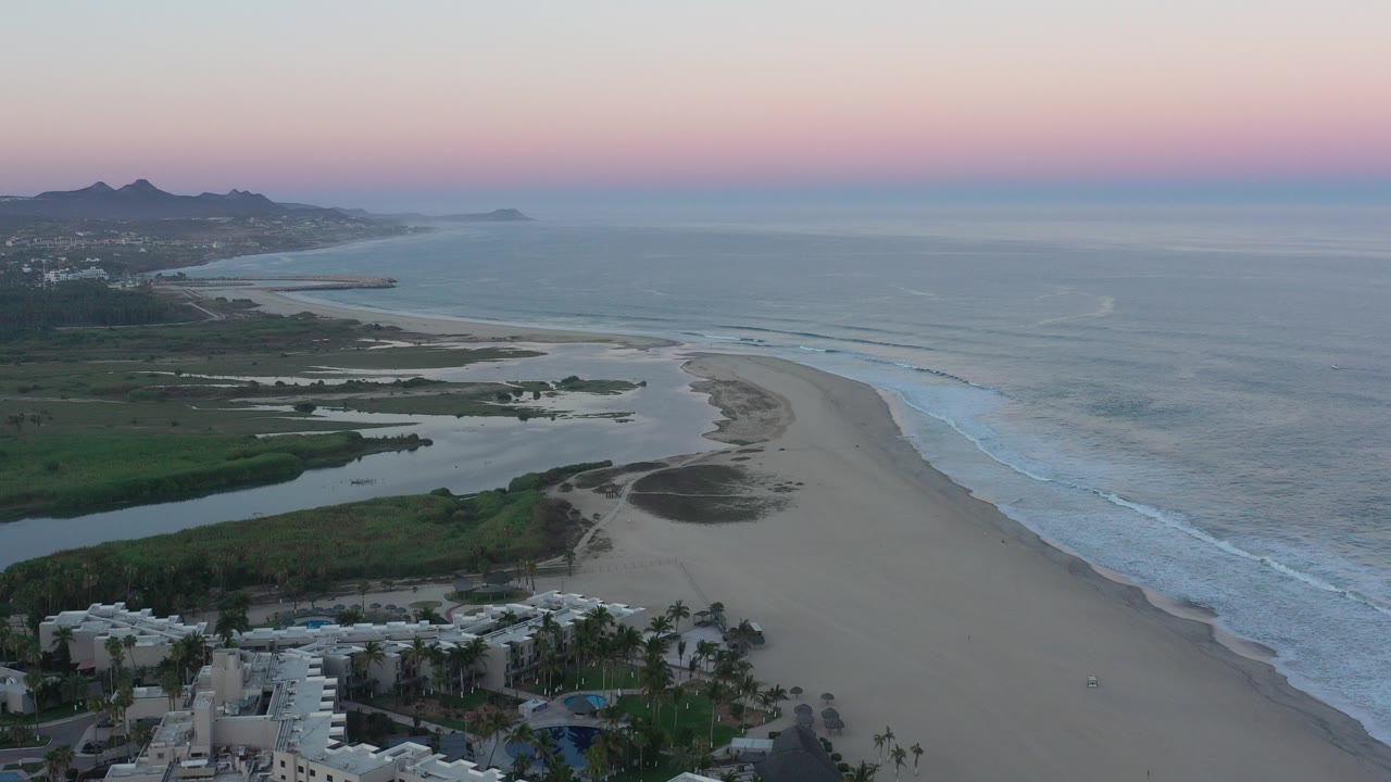 Opening aerial frame over San Jose del Cabo, Mexico, showing a curving beach beside the ocean, green lagoon areas, white buildings in the lower edge, distant hills, and no visible people.
