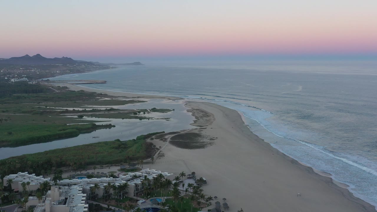 Wide aerial coastline with lagoon, beach, ocean waves, distant hills, and pink horizon.