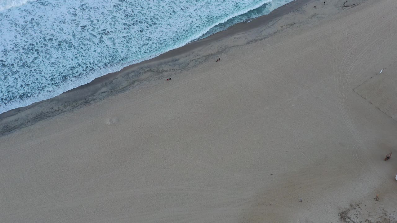 Overhead view of a San Jose del Cabo beach at the start of the clip, with foamy surf crossing the top of the frame and pale sand below.