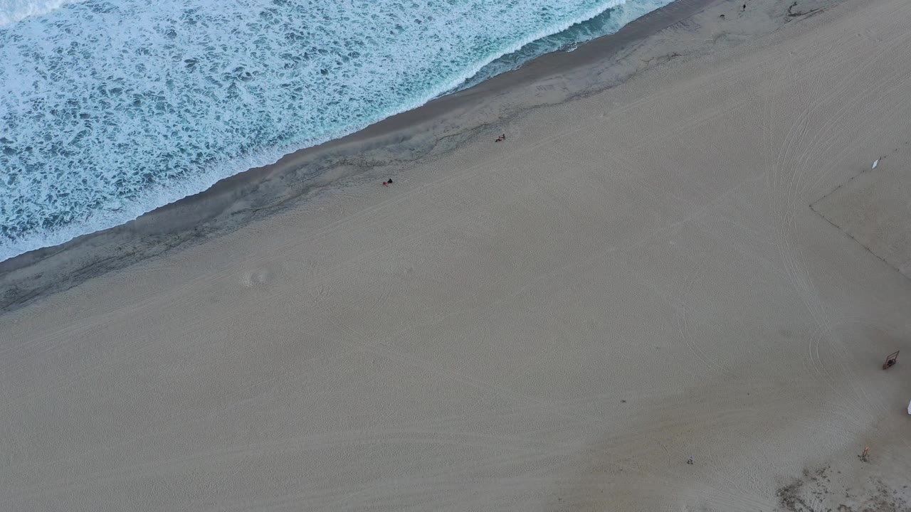 A high overhead frame shows blue-white surf advancing along the upper-left shoreline of a San Jose del Cabo beach, with open gray-tan sand below.