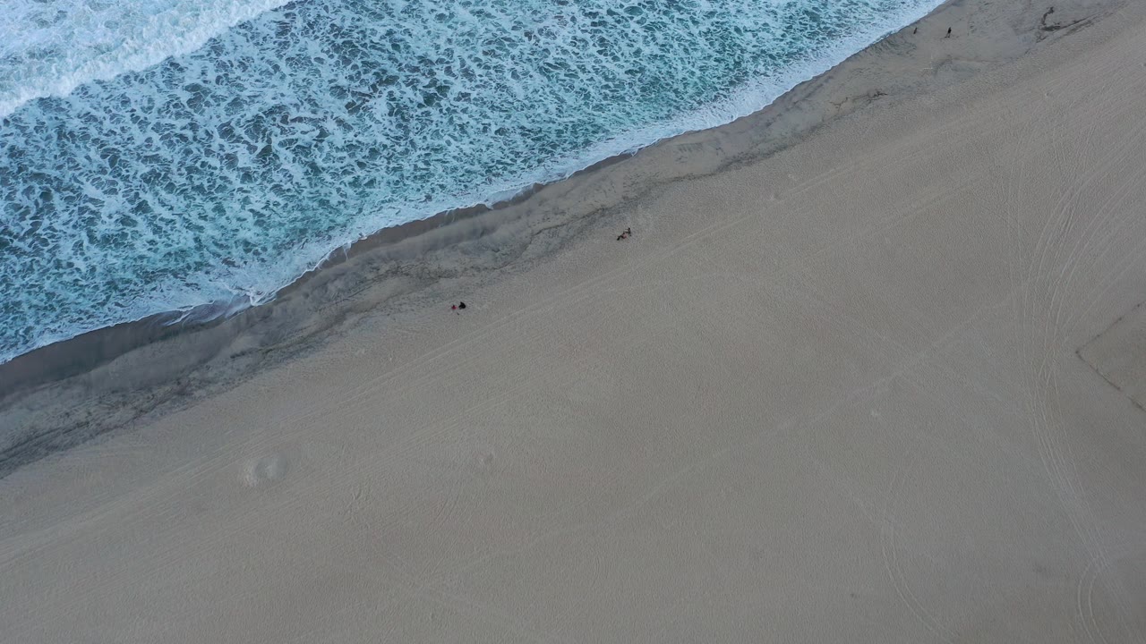 Overhead aerial frame in San Jose Del Cabo, Mexico, with foamy blue surf entering from the upper left and a broad gray-tan beach below, no visible people.