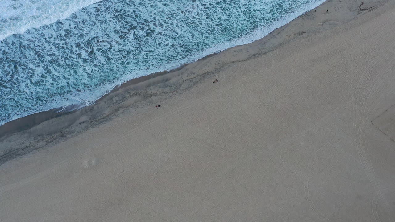 Aerial view in San Jose Del Cabo, Mexico, where white surf runs diagonally across the top of the frame and empty sand spreads through the lower right, no visible people.
