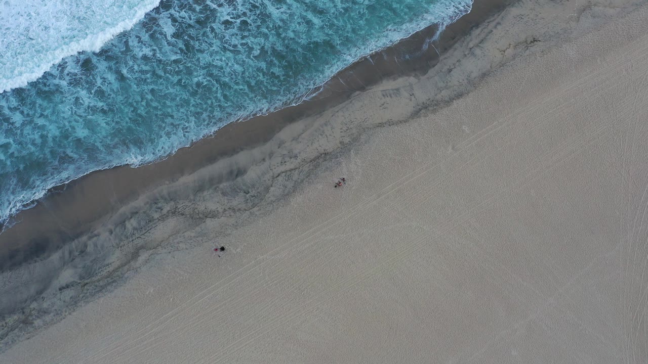 Overhead surf washing onto a sandy beach with small people visible.