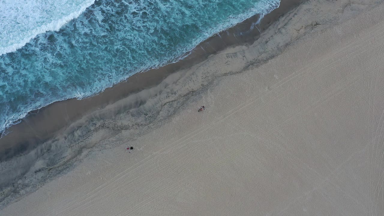 Aerial frame from San Jose del Cabo, Mexico, showing foamy water along the upper left edge, darker wet sand through the center, and small people set back on the sand.