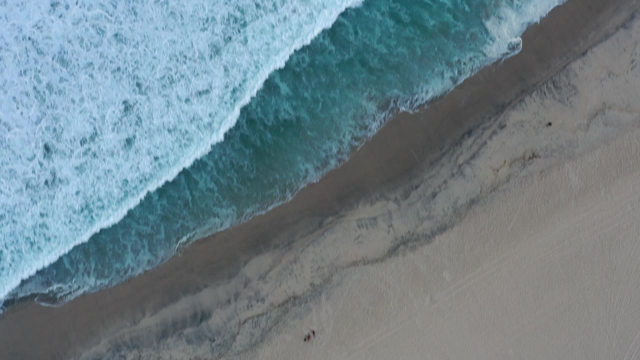 Overhead aerial frame of San Jose Del Cabo surf washing diagonally onto wet and dry sand, with teal water at the top and no visible people.