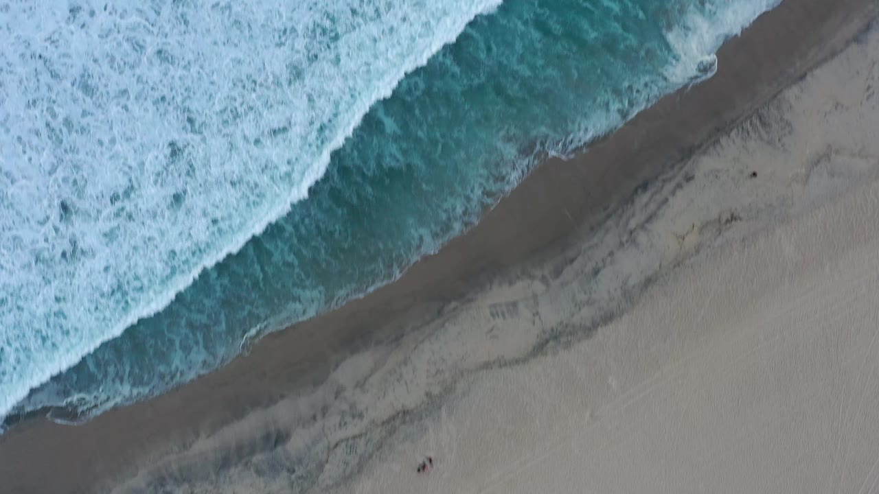 Aerial view of San Jose Del Cabo waves pressing closer across the upper frame, with a narrow darker band of wet sand below and no visible people.