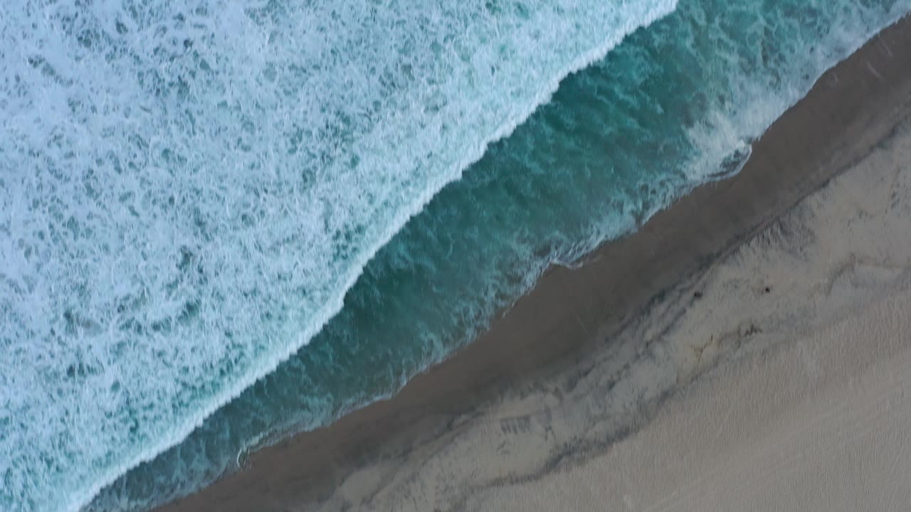 White ocean foam spreads across teal water near sand.
