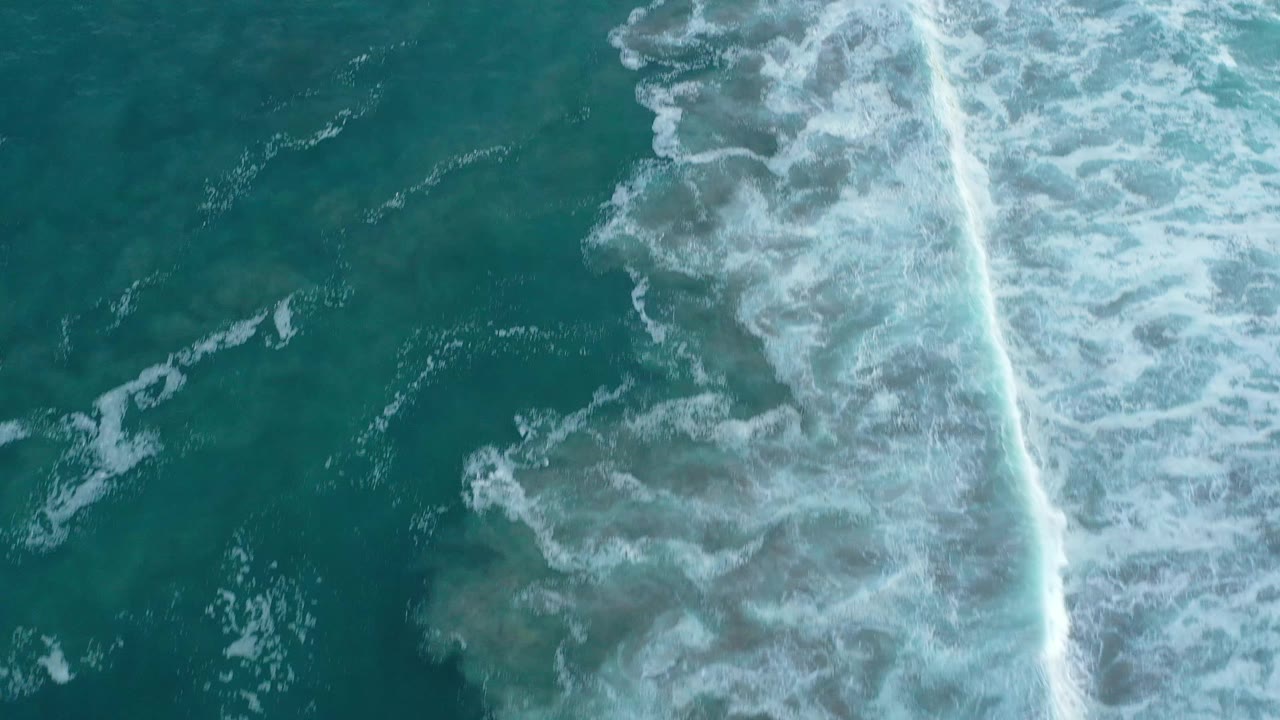 Aerial view over San Jose del Cabo water as white foam spreads along a diagonal surf band, with no visible people.