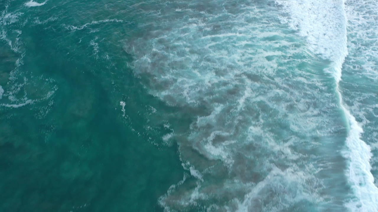 Aerial top-down frame over San Jose del Cabo ocean water, where a broad white foam band runs down the right side against green-blue water, no visible people.