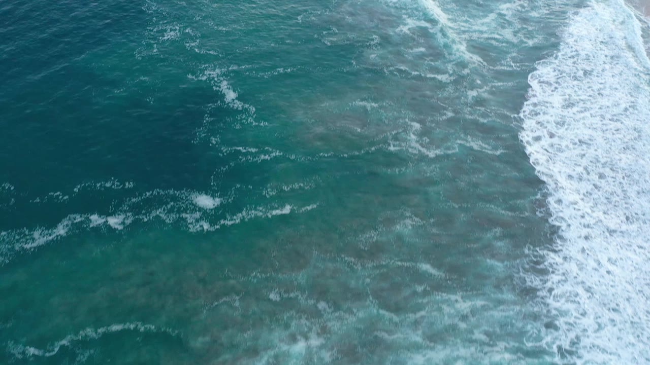 Aerial frame of white wave foam pushing farther into teal-green water near the shoreline in San Jose del Cabo, Mexico, with no visible people.