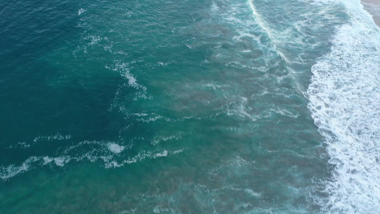 Overhead ocean surface with broken foam trails and shoreline surf.