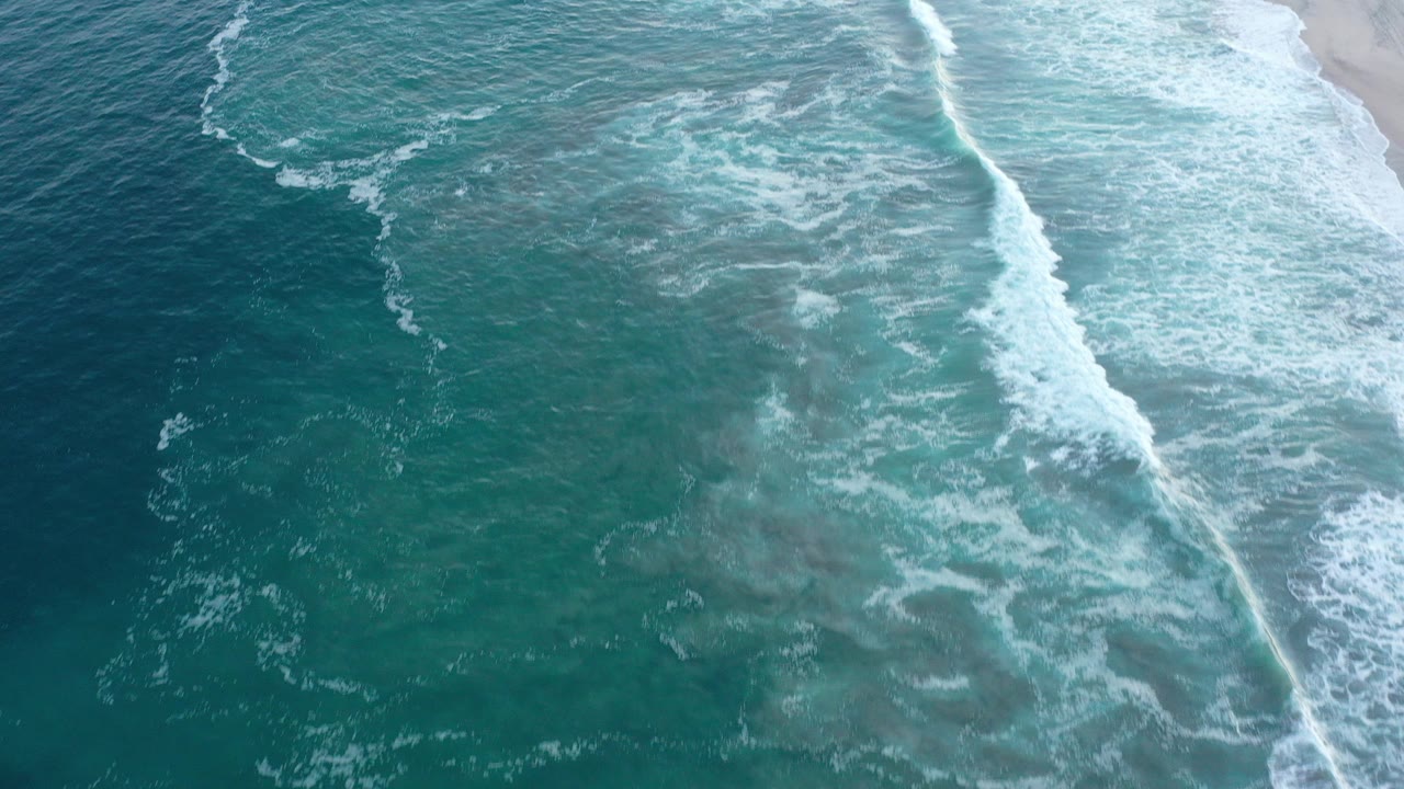 Aerial frame over San Jose del Cabo surf, with turquoise water and a diagonal white wave line reaching pale sand on the right, no visible people.