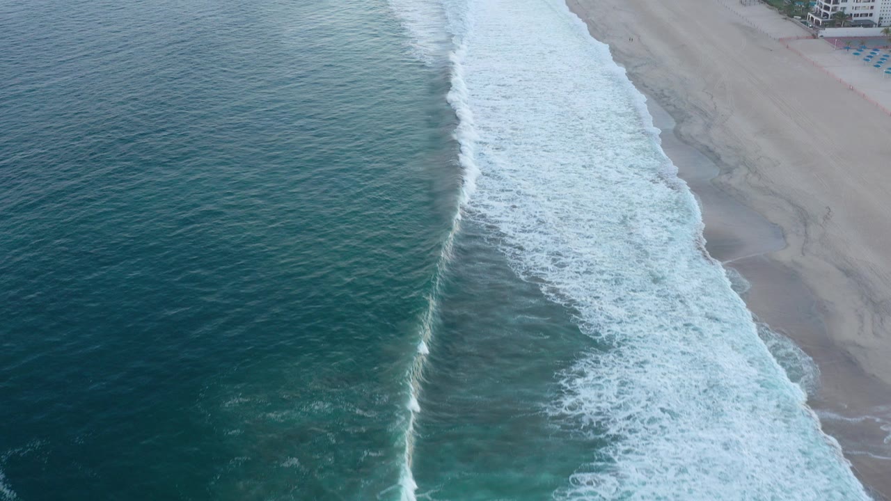Aerial shoreline with teal water, white surf, pale sand, and distant beachfront buildings.