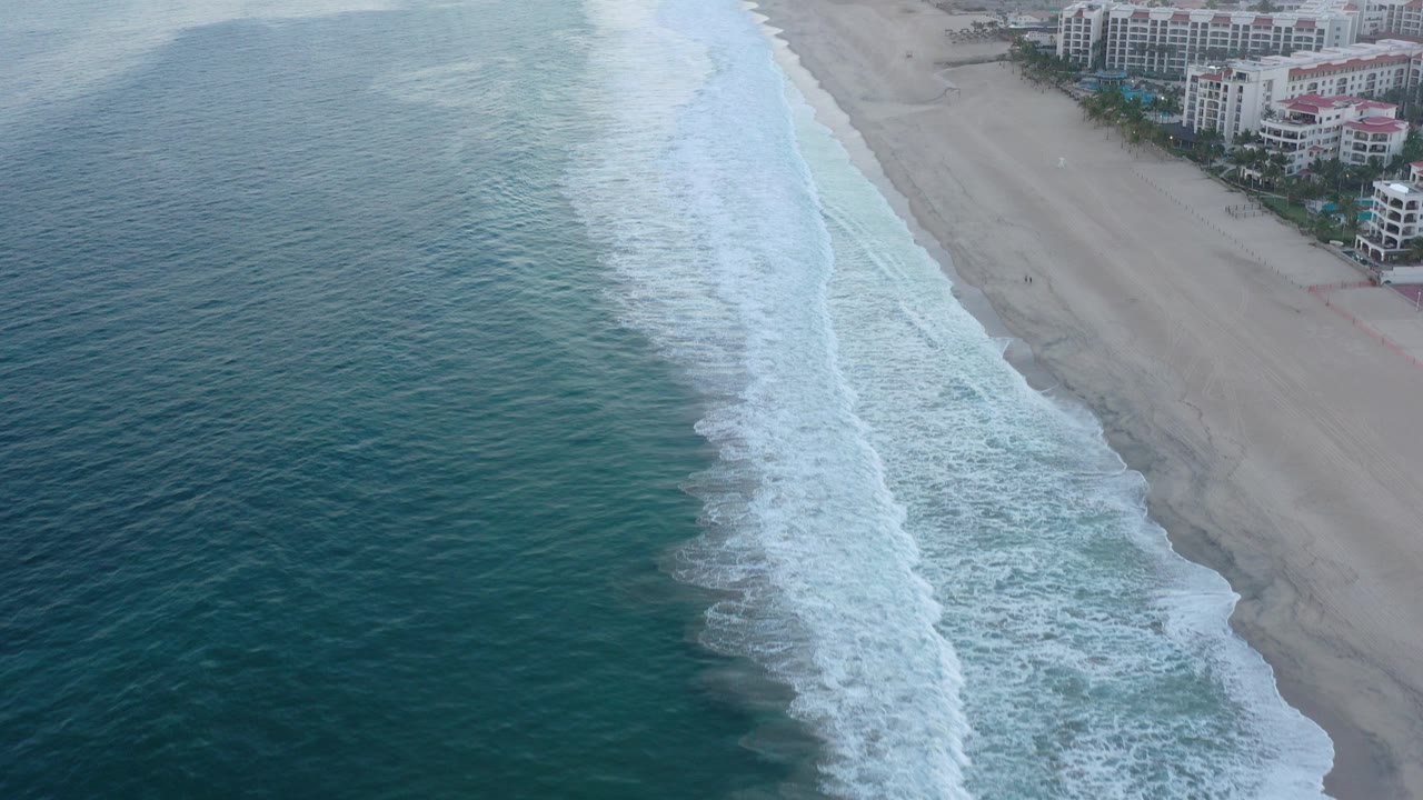 High aerial view over San Jose del Cabo, Mexico with teal ocean on the left, white surf bands along the sand, beachfront hotels at the upper right, and no visible people.