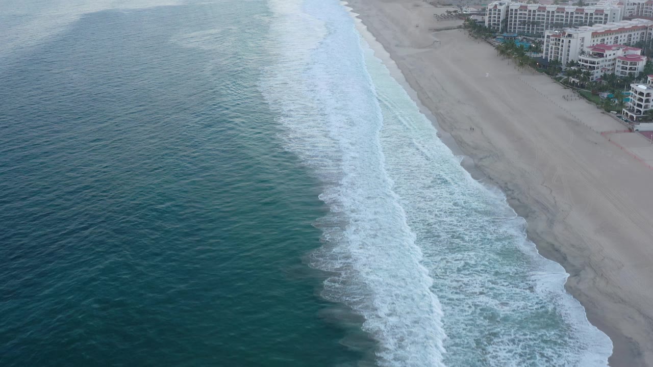 Aerial frame in San Jose del Cabo, Mexico showing a longer diagonal band of white surf between darker ocean water and pale sand, with hotels near the top edge and no visible people.