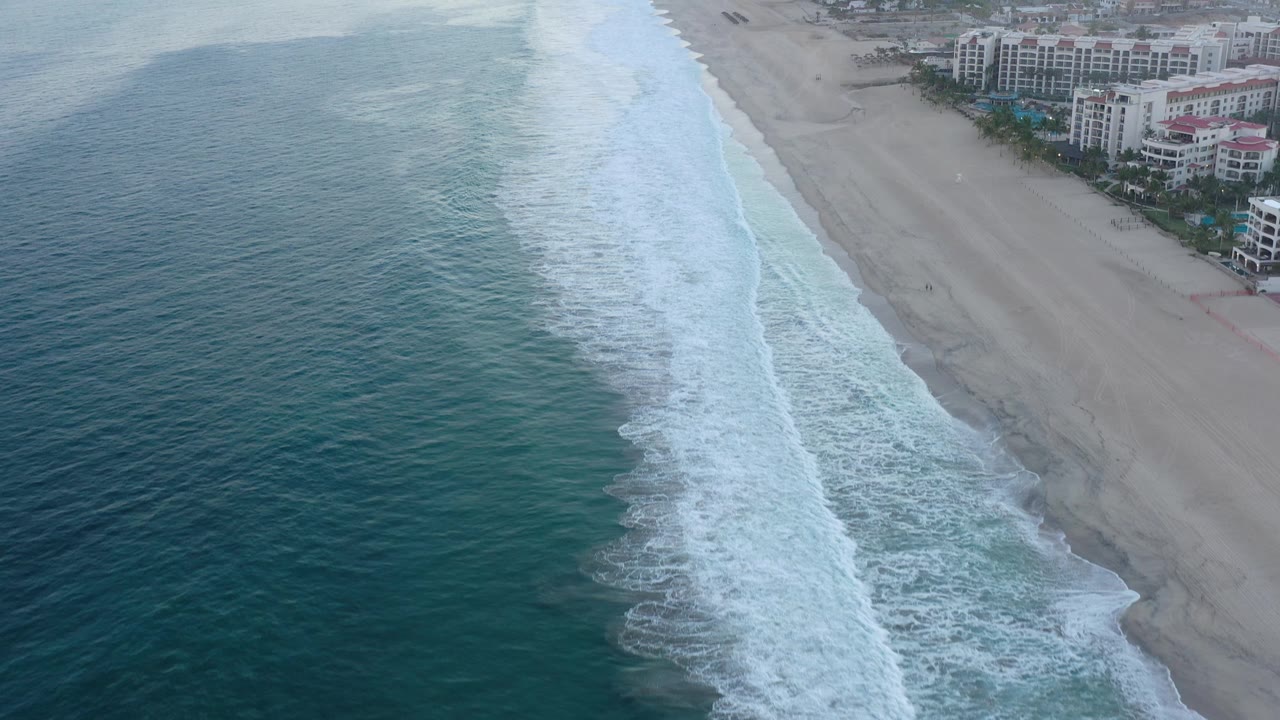 White waves roll toward a broad beach with resort buildings beyond the sand.