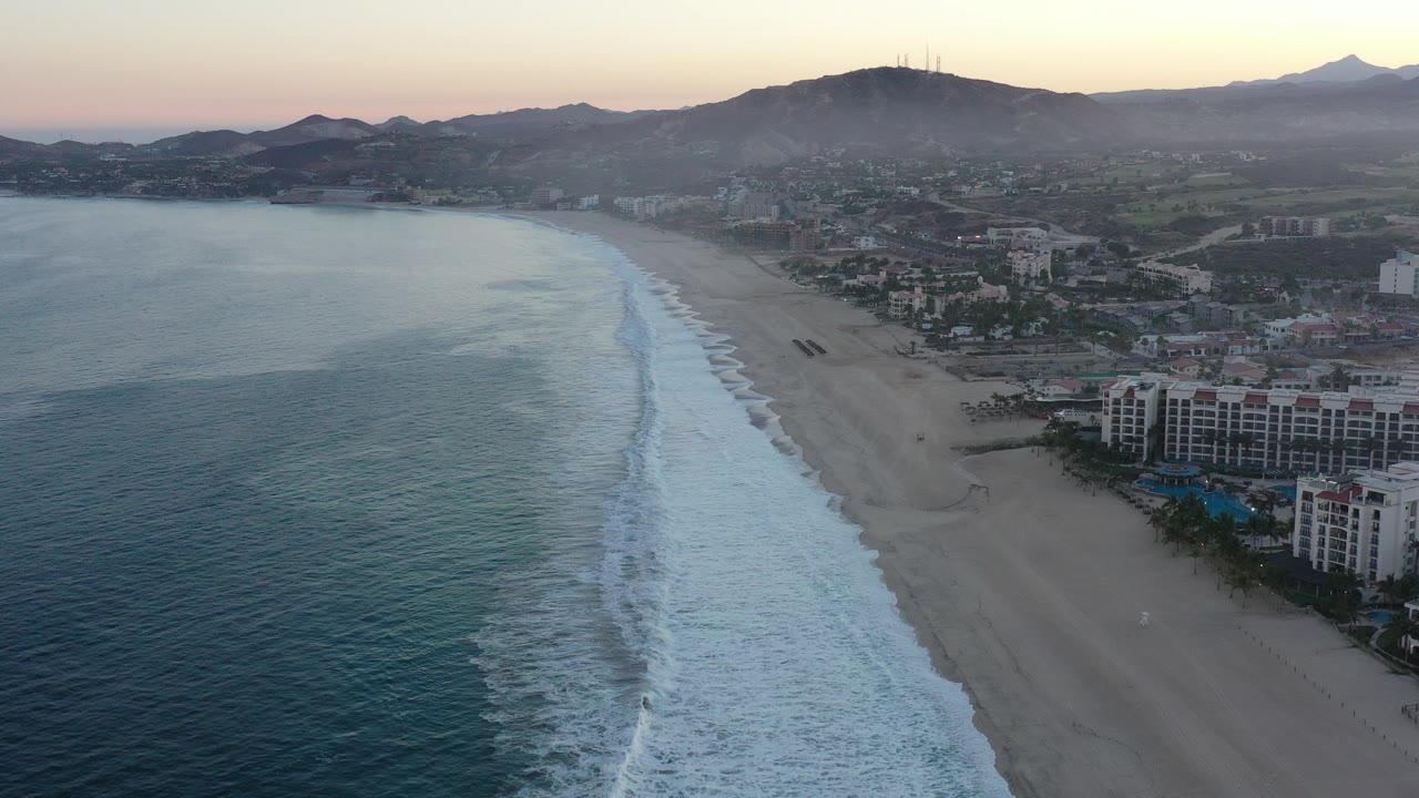 Aerial view of surf, beach, resort buildings, town, and mountains at dusk.