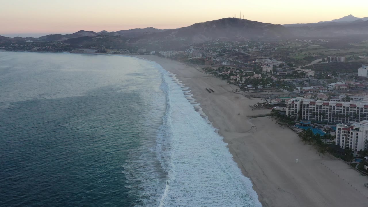 A second aerial frame of San Jose del Cabo shows the surf band slightly farther along the beach, with hotel buildings near the lower shore, inland streets, mountains, and no visible people.