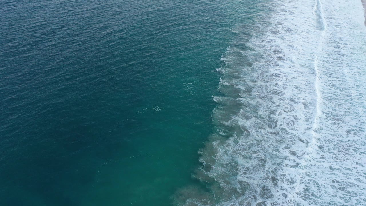 A band of white foam advances through blue-green ocean water beside the San Jose del Cabo beach edge, with no visible people.