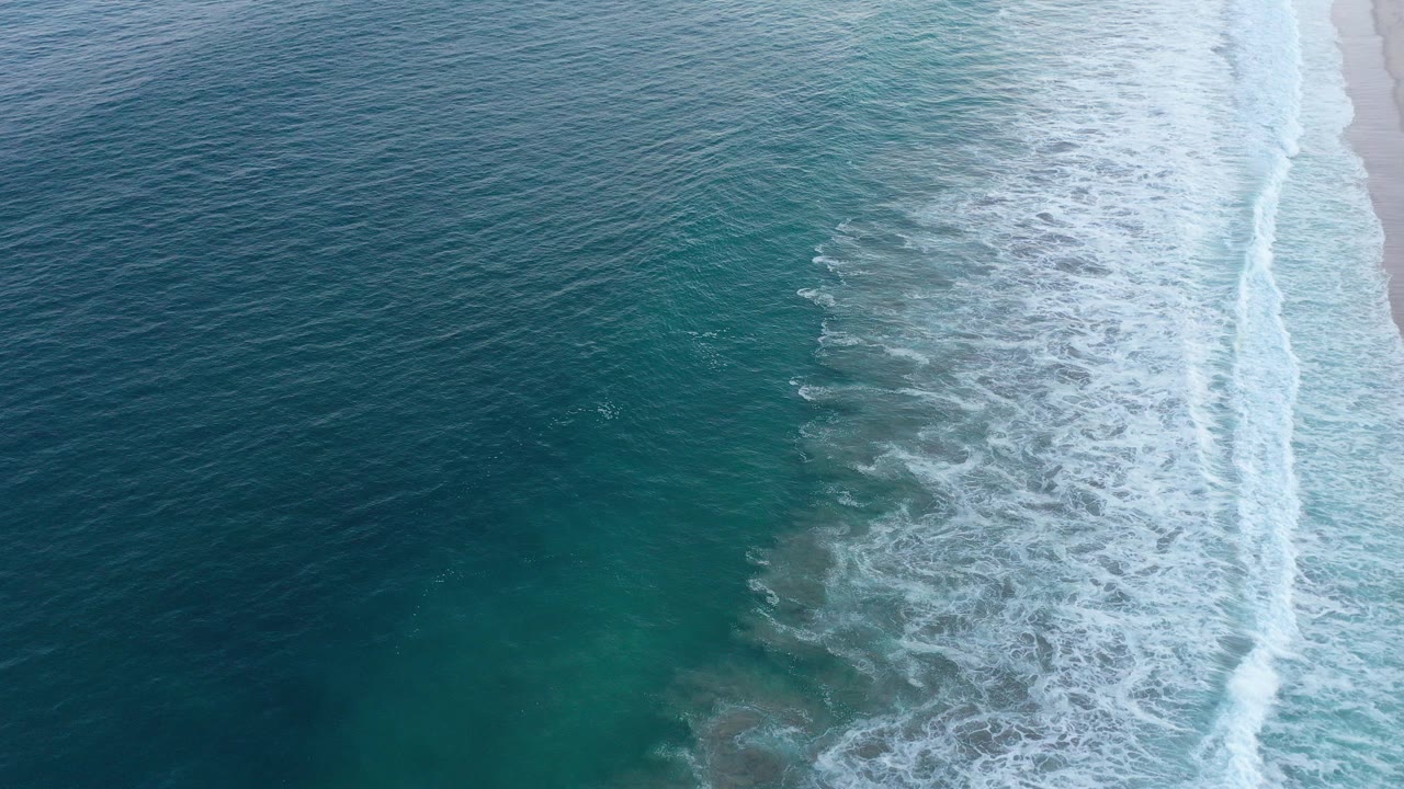 Aerial view over San Jose Del Cabo, Mexico, with dark blue water on the left, white surf crossing toward pale sand on the right, and no visible people.