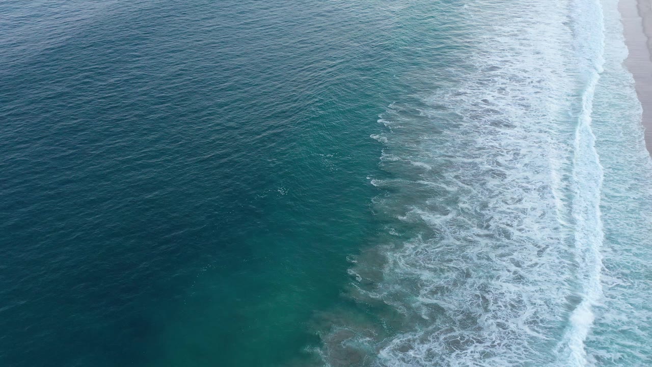 Aerial San Jose Del Cabo shoreline frame with foamy waves widened across the center, deeper blue sea at left, a narrow strip of sand at right, and no visible people.