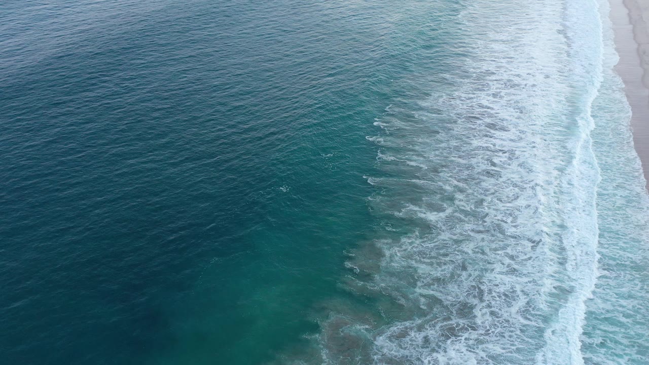 Top-down shoreline with blue water, white breakers, and sand.
