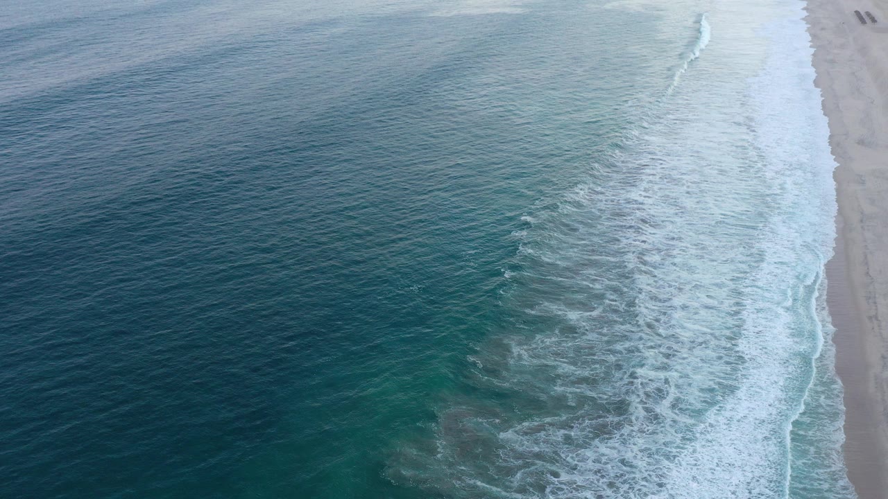 Aerial view over San Jose Del Cabo water, with blue-green ocean on the left, white foam bands near the beach on the right, and no visible people.