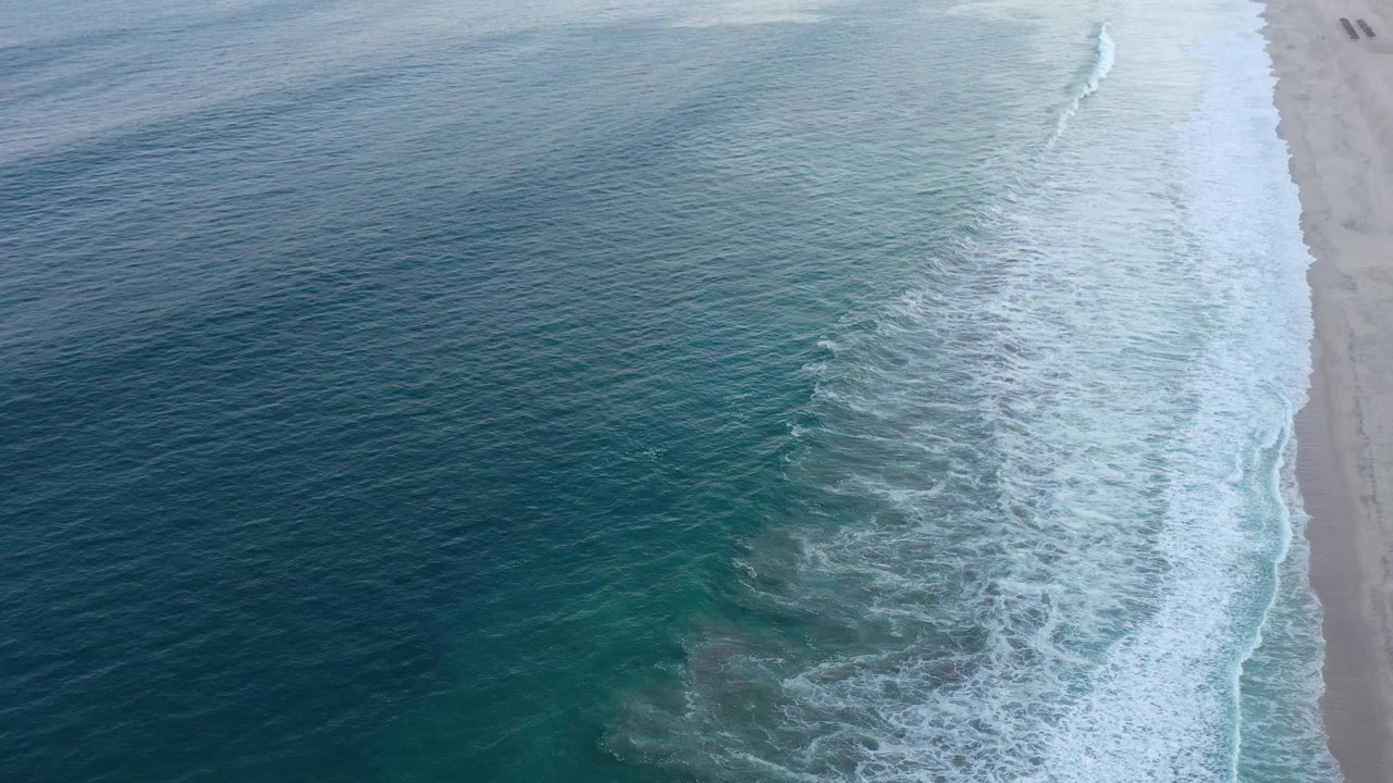 Drone-height view of the San Jose Del Cabo shoreline, with textured blue water, a wide white surf strip, pale sand at the right edge, and no visible people.